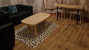 Una sala de estar con piso de madera, un sofá negro con un cojín azul, una mesita y dos sillas. Hay una araña de techo en el techo.