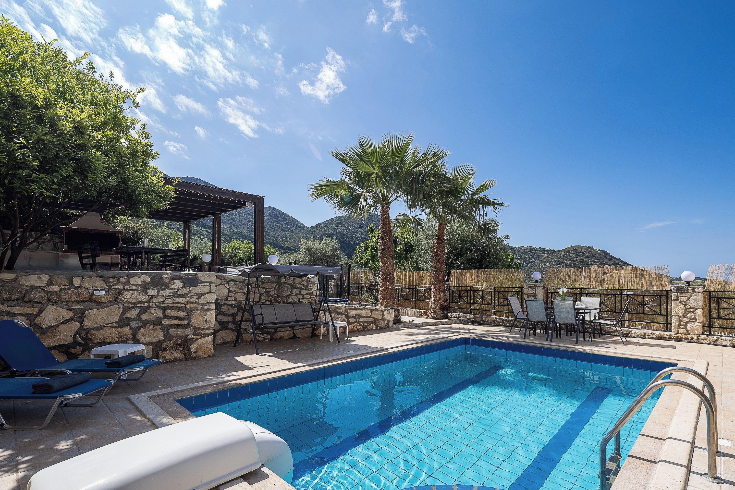 Una piscina con lettini e un muro in pietra, circondata da palme e uno sfondo montuoso.