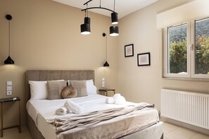 A neatly made bed with a beige headboard, a bedside table with a lamp, and a window with a view of greenery.