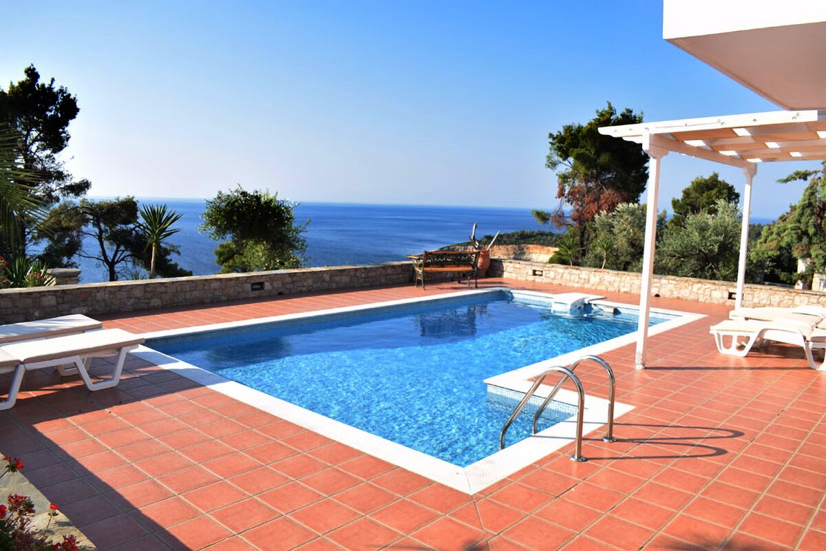 A swimming pool with a view of the sea, surrounded by lounge chairs and a pergola.