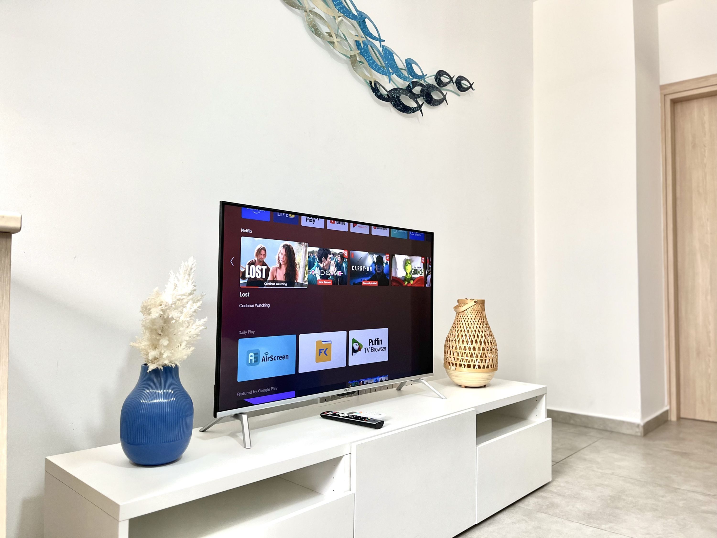 A flat-screen TV on a white entertainment unit with a remote control, a blue vase, and a decorative wall piece.