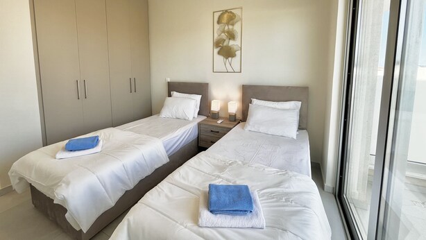 A hotel room with two beds, a wardrobe, bedside lamps, and a window. - 85 KAP - Stay Mythical Blue Apartment (Paralimni)