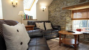 A cozy living room with a stone wall, a leather sofa, a wooden coffee table, and a bookshelf. - Bluebell Cottage, Aberporth (Aberporth)
