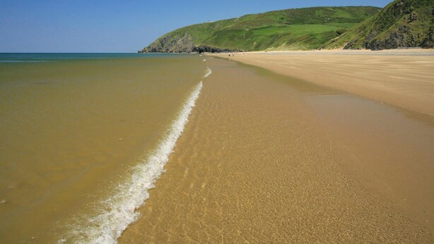 A sandy beach with gentle waves, a clear sky, and a green hillside in the background. - Bluebell Cottage, Aberporth (Aberporth)