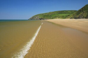 A sandy beach with gentle waves, a clear sky, and a green hillside in the background. - Bluebell Cottage, Aberporth (Aberporth)