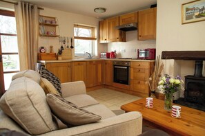 A cozy living room with a sofa, a wooden coffee table with mugs, a kitchen with wooden cabinets, a stove, and a window with curtains.