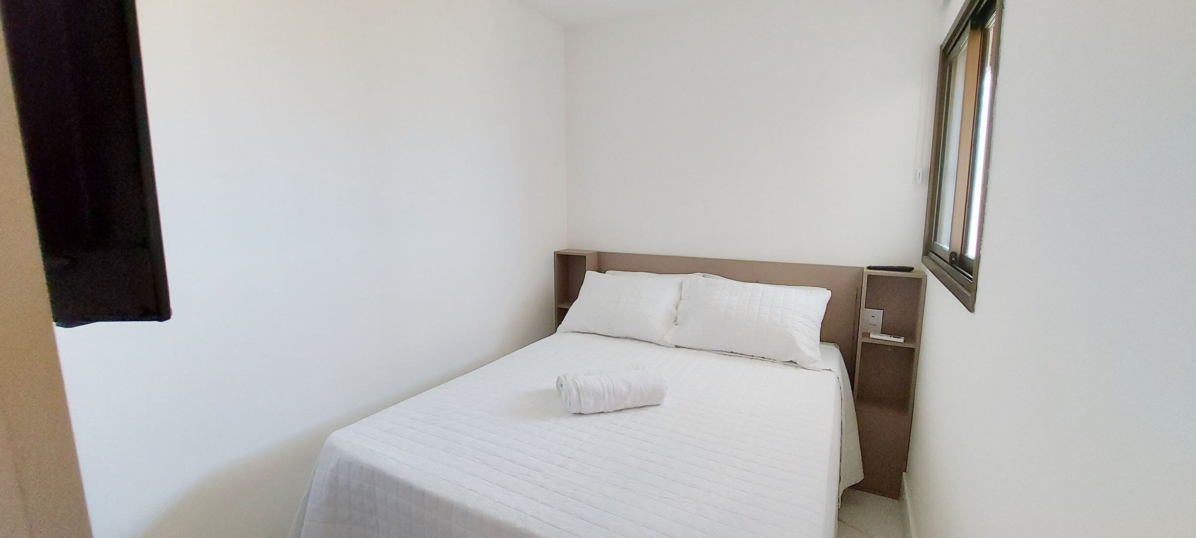 A small, neatly made bed with white linens in a corner room with white walls and a wooden headboard.