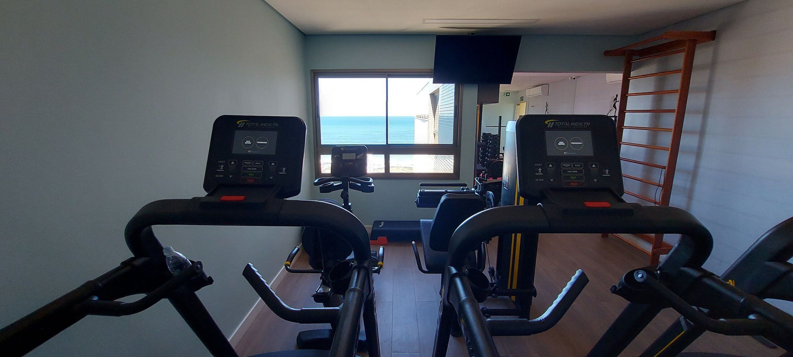 Ein Heimfitnessraum mit zwei Ergometer, einem an der Wand befestigten Fernseher und einem Fenster mit Meerblick.