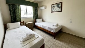 A hotel room with two beds, a small wooden nightstand, a window with green curtains, an air conditioning unit, and a framed picture on the wall. - 101 AN -chill n Joy Apartment (Ayia Napa)