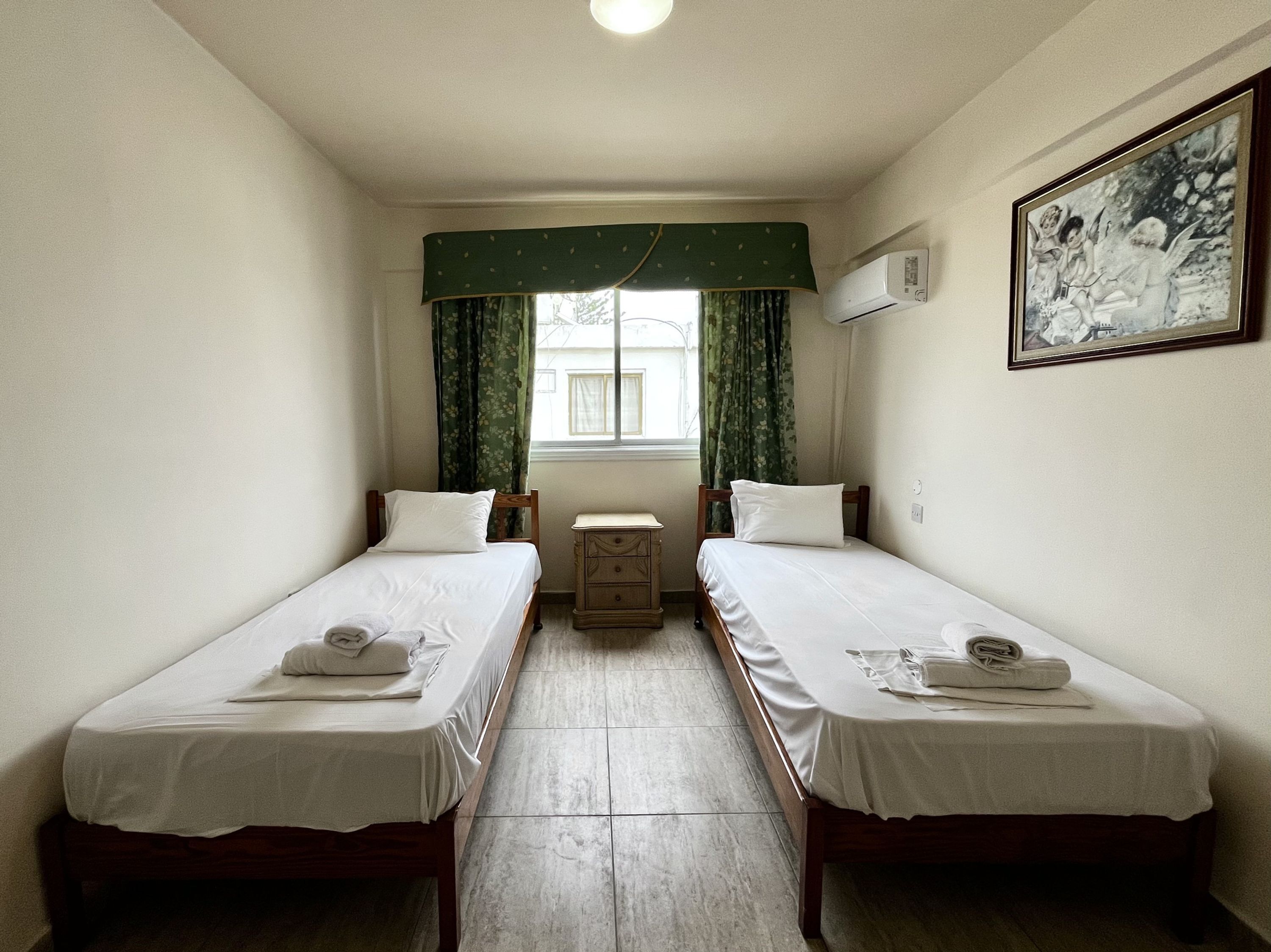A room with two single beds, a small dresser, a window with curtains, and a framed picture on the wall.