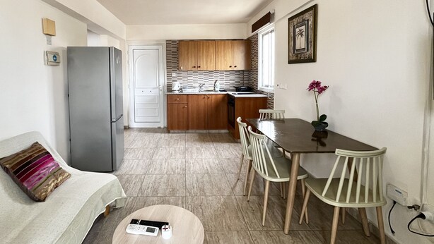 A compact kitchen with wooden cabinets, a dining table with chairs, a gray refrigerator, and a small round table with a remote control. - 101 AN -chill n Joy Apartment (Ayia Napa)