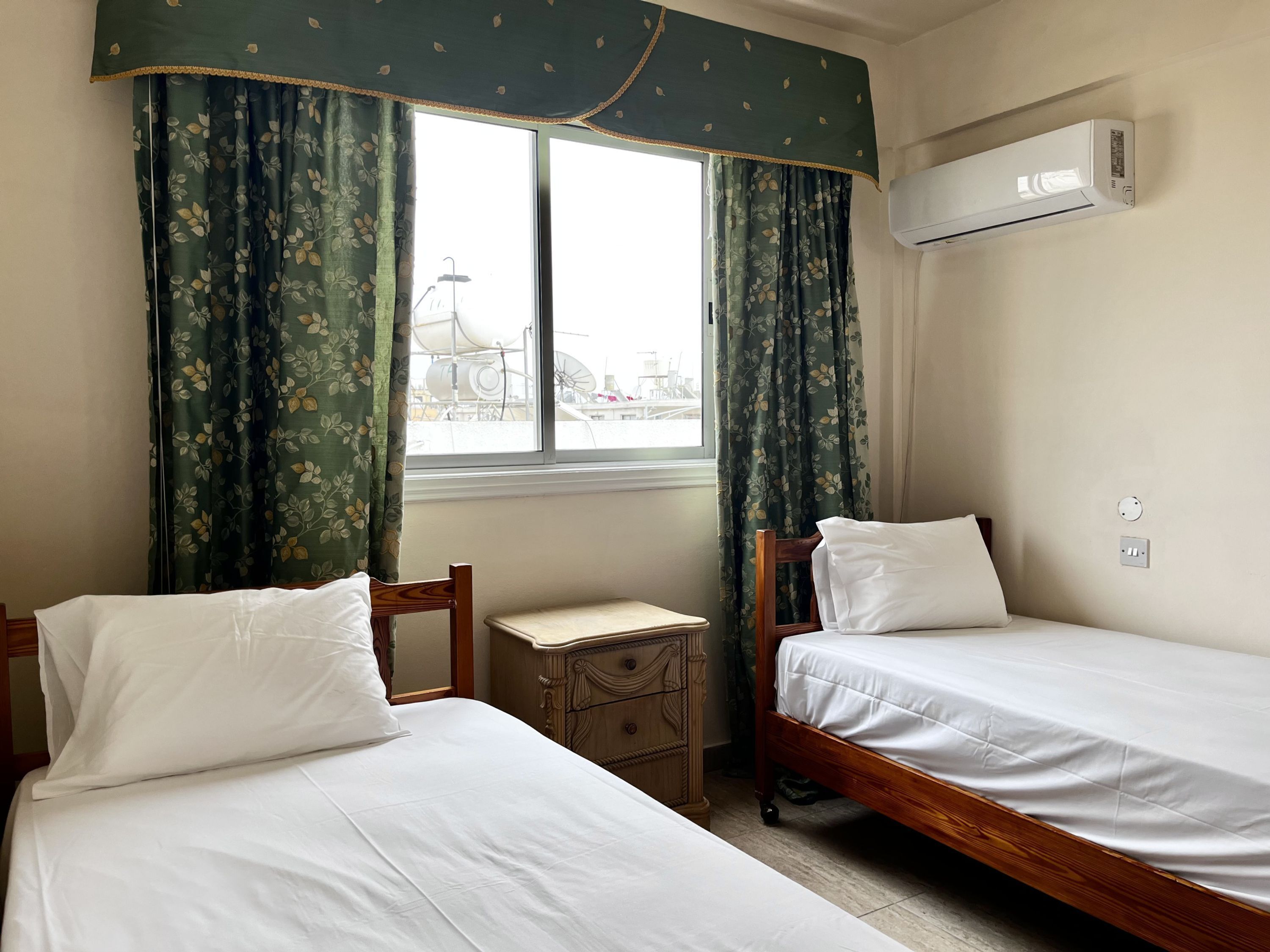A room with two single beds, a small wooden nightstand, a window with curtains, and an air conditioning unit.