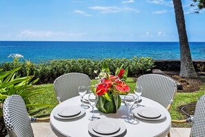 Une table ronde dressée pour quatre personnes, avec de l’argenterie blanche et des verres, un vase rempli de fleurs rouges et une vue sur l’océan.