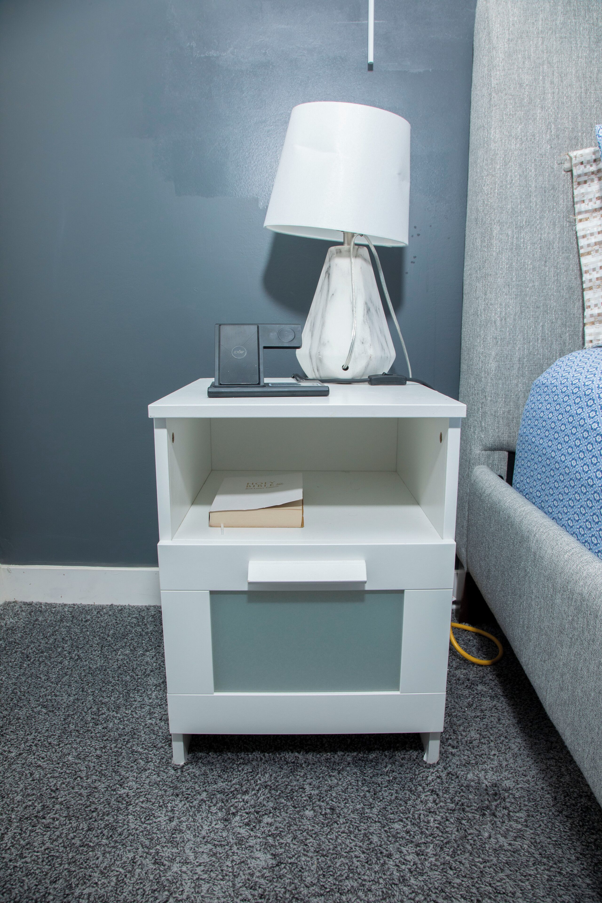 A white nightstand with a lamp, a book, and a small electronic device.