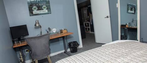 Un dormitorio con una cama, un escritorio con computadora, una silla y una puerta que da a otra habitación.
