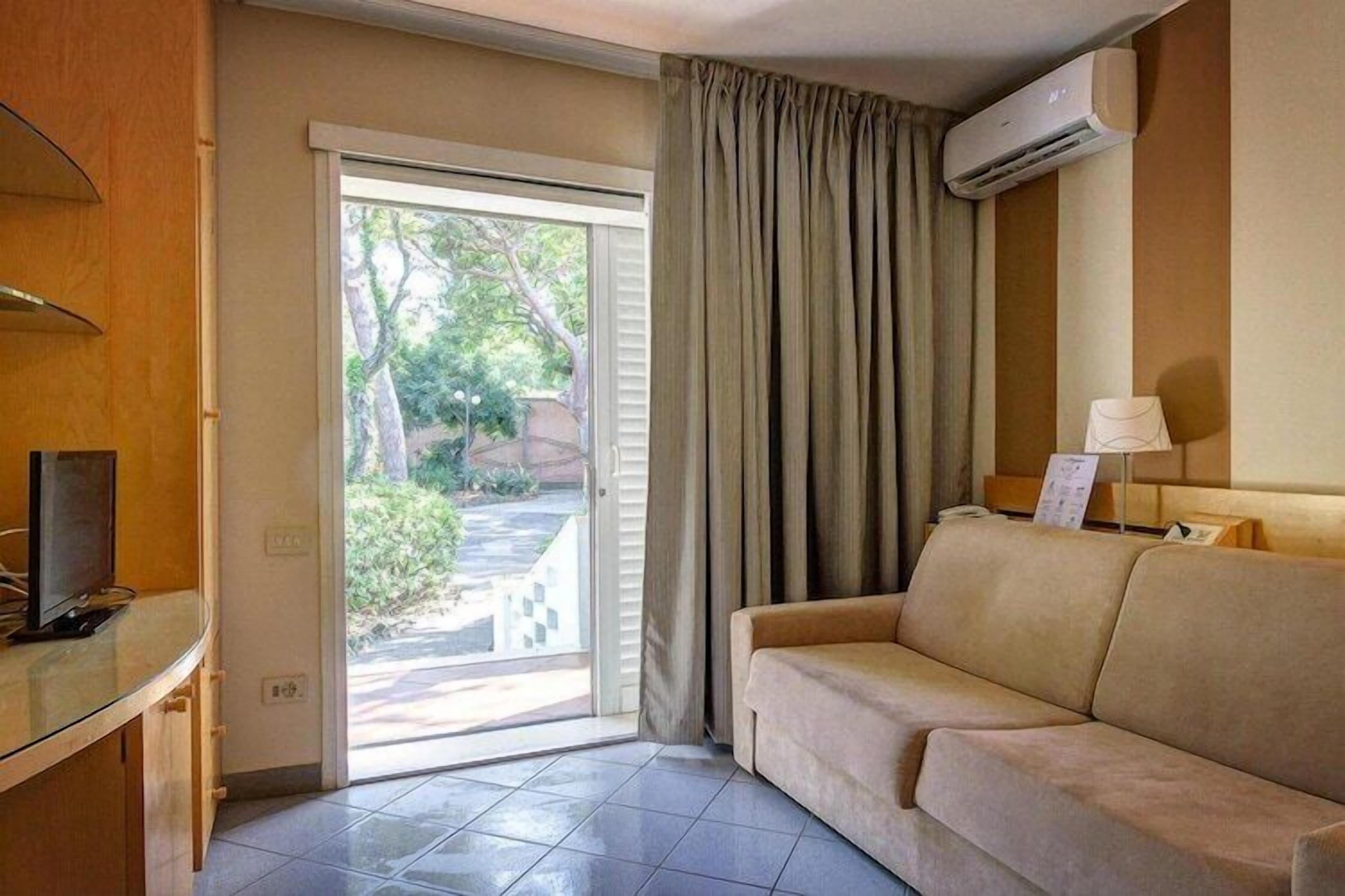 A hotel room with a sofa, a TV on a stand, a sliding glass door leading to a garden, and an air conditioning unit on the wall.