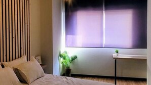 Une chambre avec un grand lit, un luminaire au plafond, une fenêtre avec des stores et une petite table sur laquelle est posée une plante.