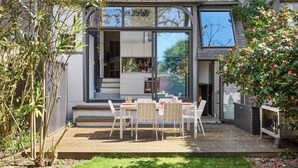 Una casa moderna con un patio, un comedor con sillas blancas y un jardín con diversas plantas.
