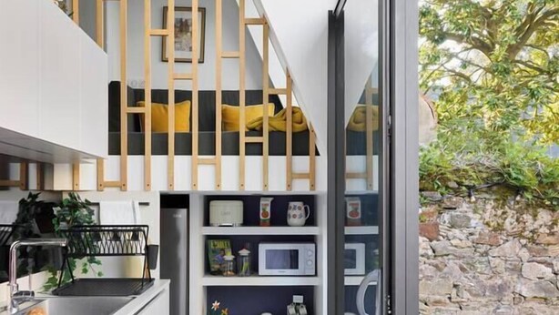 Una cocina compacta con estufa integrada, microondas y un refrigerador pequeño. Una escalera de madera conduce a una zona estilo altillo con una cama.
