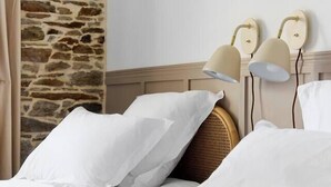 Habitación de hotel con pared de piedra, un espejo redondo, dos lámparas de noche y una cama con ropa blanca.
