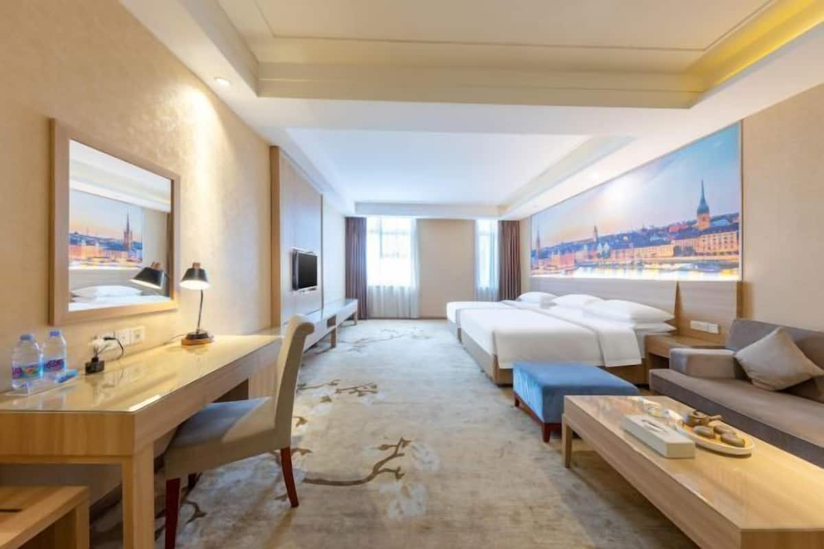 A hotel room with a bed, a desk, a chair, a sofa, a TV, and a painting of a cityscape.