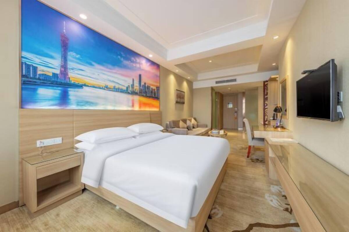 A hotel room with a bed, a TV, a painting of a cityscape, and a desk with a chair.