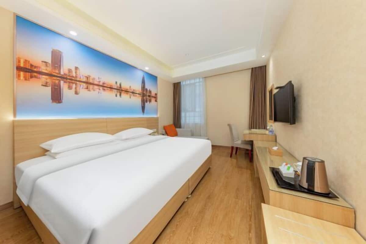 A hotel room with a large bed, a desk, a chair, a flat-screen TV, and a cityscape mural on the wall.