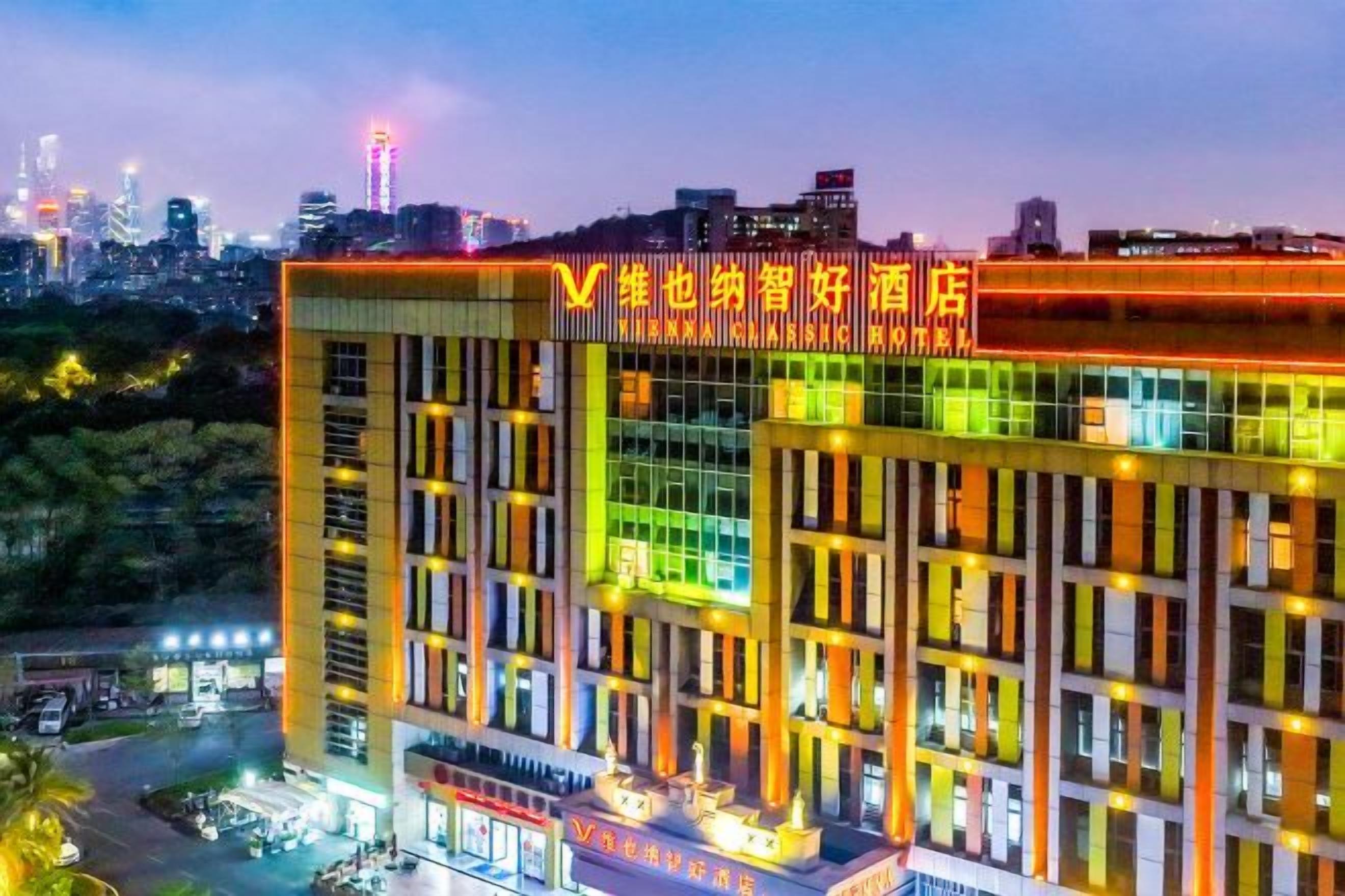 A multi-story hotel with a modern facade, illuminated signage, and a city skyline in the background.