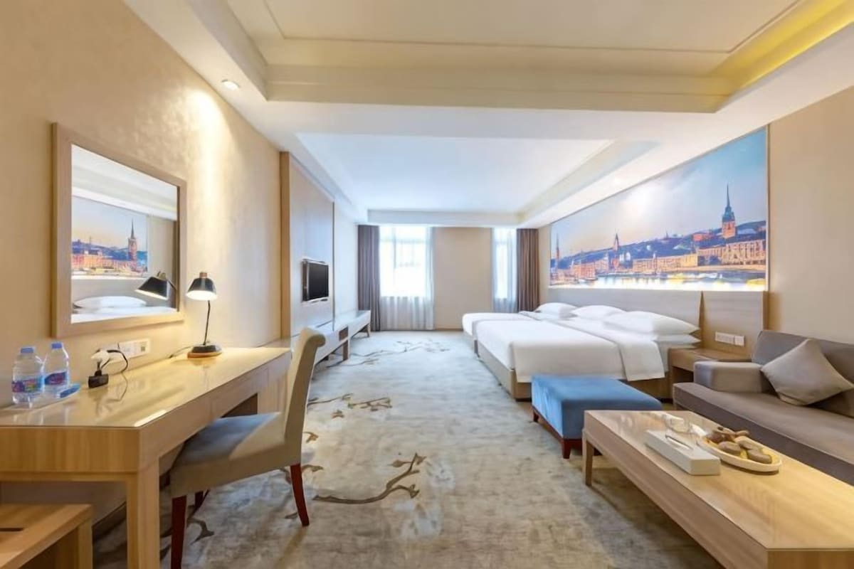 A hotel room with a bed, a desk, a chair, a sofa, a TV, and a painting of a cityscape.