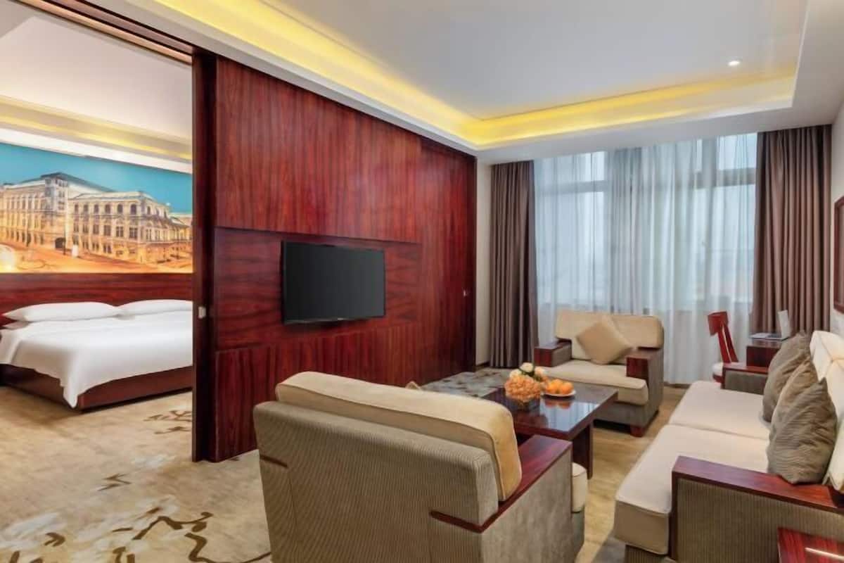 A hotel room with a bed, a sofa, a TV, and a painting of a building on the wall.