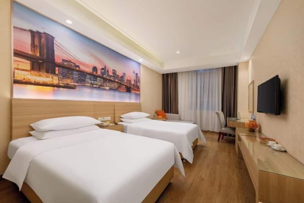A hotel room with two beds, a desk, a TV, and a cityscape mural on the wall.
