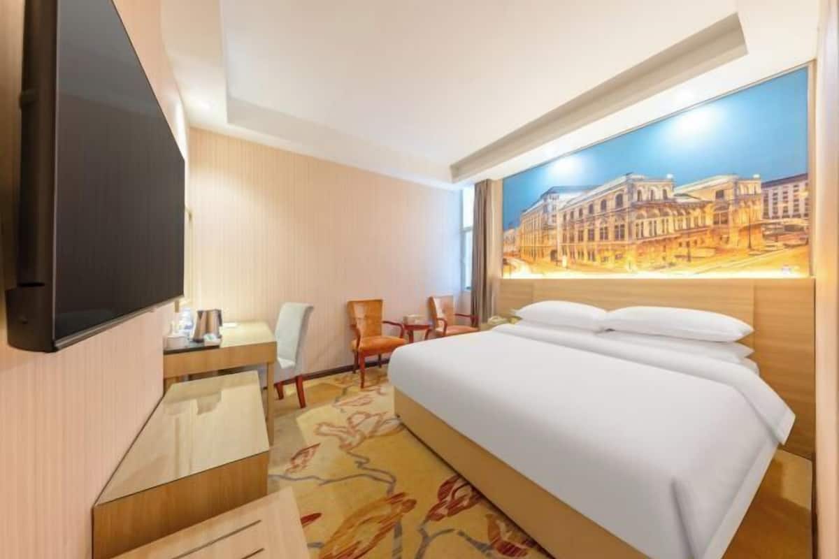 A hotel room with a bed, a desk, a chair, a television, and a painting of a cityscape.
