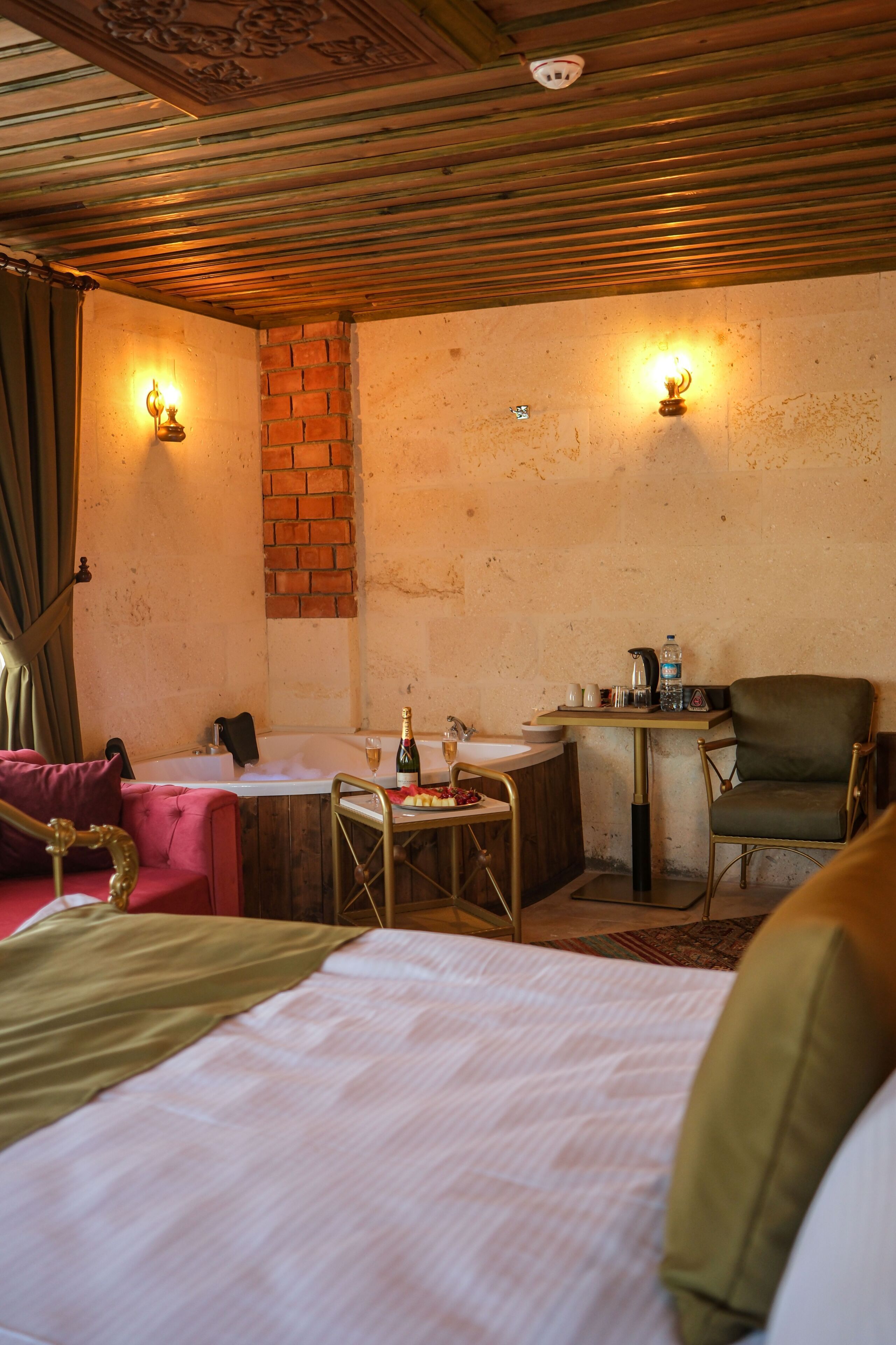 Une pièce avec un lit, une petite table sur laquelle se trouvent une bouteille et des verres, une chaise et un mur en briques agrémenté de deux lampes fixées au mur.