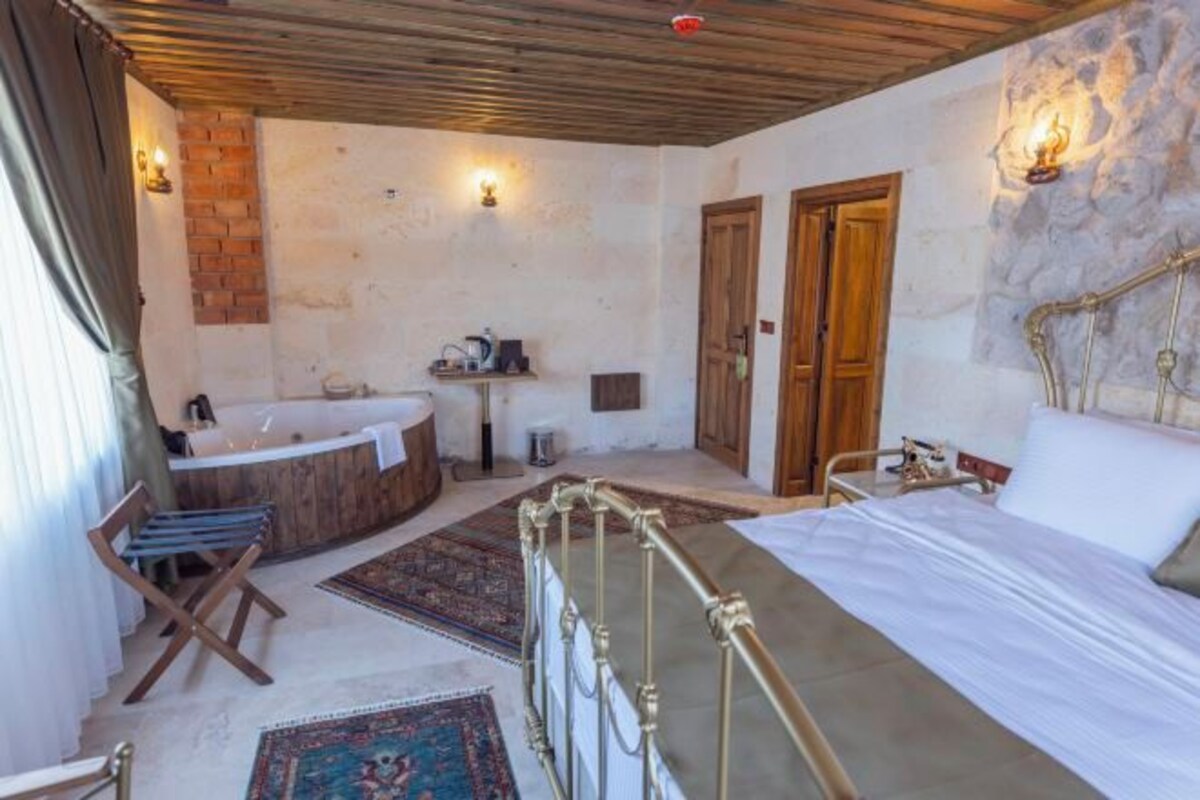 A bedroom with a bed, a wooden headboard, a small table, a chair, a bathtub, and a stone wall.