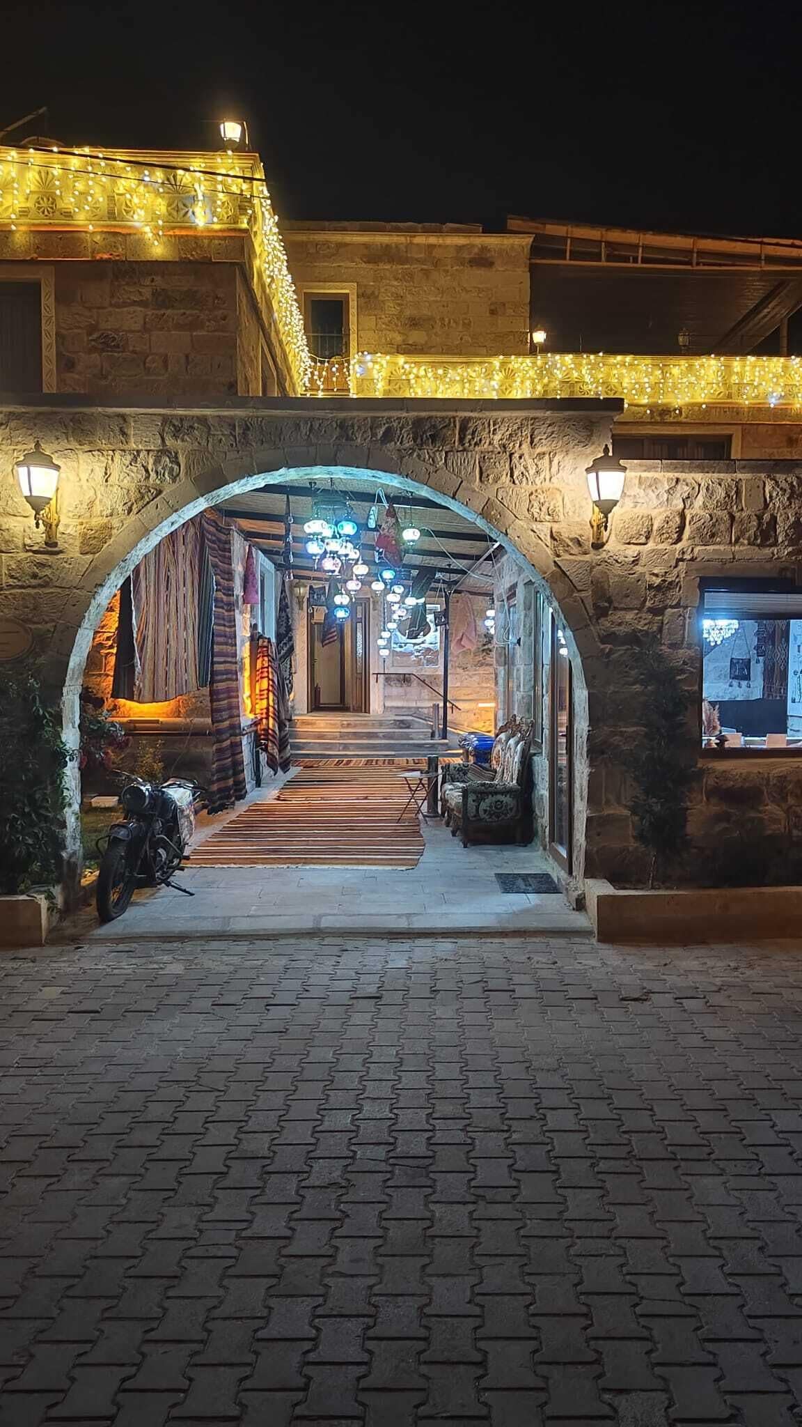 Une entrée en arc, avec un chemin pavé de briques menant à un bâtiment, illuminé par des guirlandes lumineuses et des lampes fixées aux murs.