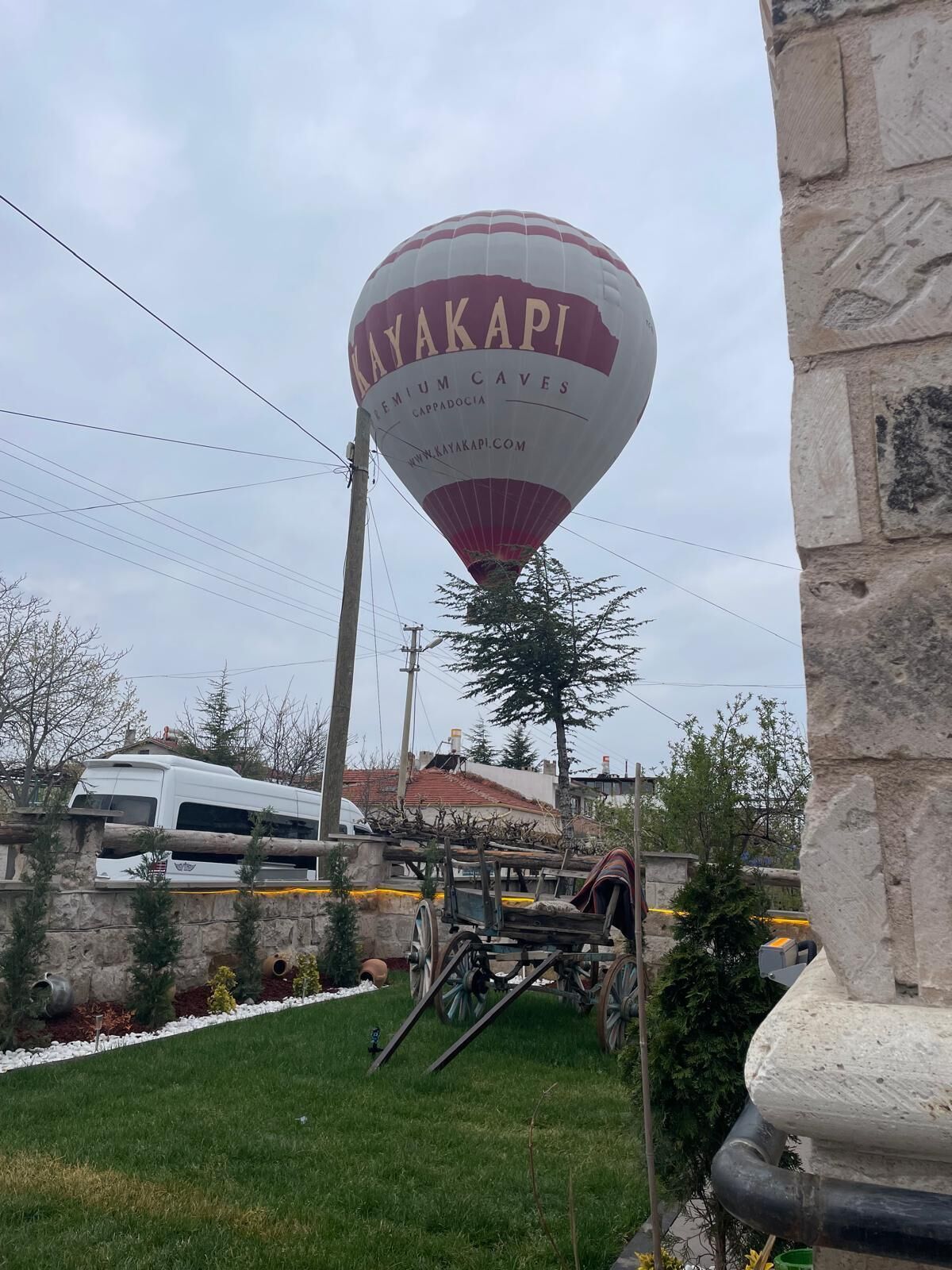 Un globo aerostático con el texto 'KAYAKAPI' y 'Cuevas Premium', un muro de piedra, un vehículo blanco y un carrito de madera.