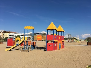 A beach playground with slides and climbing structures, set against a backdrop of buildings and clear skies. - Three-room apartment 350 m. from the sea in Bibione Pineda. (Bibione Pineda)
