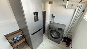 A compact kitchen with a stainless steel refrigerator, a washing machine, and a small wooden shelf with various items. - Bright 3-room apartment in the center of Royan, 3-minute walk from the beach and port. (Royan)