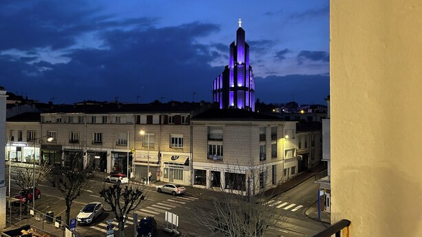 A building with a prominent tower featuring a cross at the top, surrounded by a street with parked cars and storefronts. - Bright 3-room apartment in the center of Royan, 3-minute walk from the beach and port. (Royan)