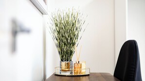 Une table à manger agrémentée d’un élément décoratif central composé d’une grande plante verte dans un pot à motifs, accompagnée de deux photophores en verre.