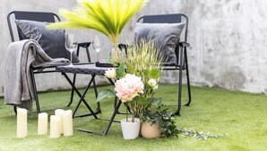 Un coin repas avec deux chaises noires, une table avec un verre, un vase de fleurs et des bougies posées sur l’herbe.
