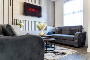 A modern living room with a grey sofa, a coffee table, and a TV mounted on the wall displaying "NETFLIX".