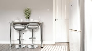 Une cuisine avec un réfrigérateur blanc, deux tabourets de bar, une petite table ornée d’un vase de plantes et une porte.