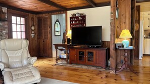 A wooden-paneled living room with a leather armchair, a television on a wooden stand, and a small table with a lamp.