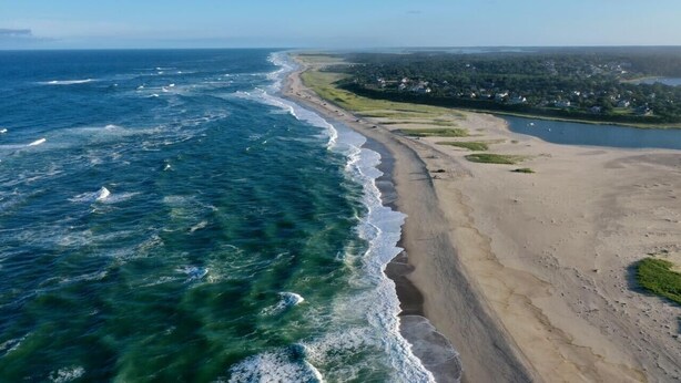 An aerial view of a coastal area with a sandy beach, a clear blue ocean, and a distant shoreline with buildings.