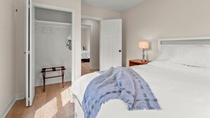 Un dormitorio con cama, una mesita de noche con lámpara, un banco de madera y un armario abierto con perchas.