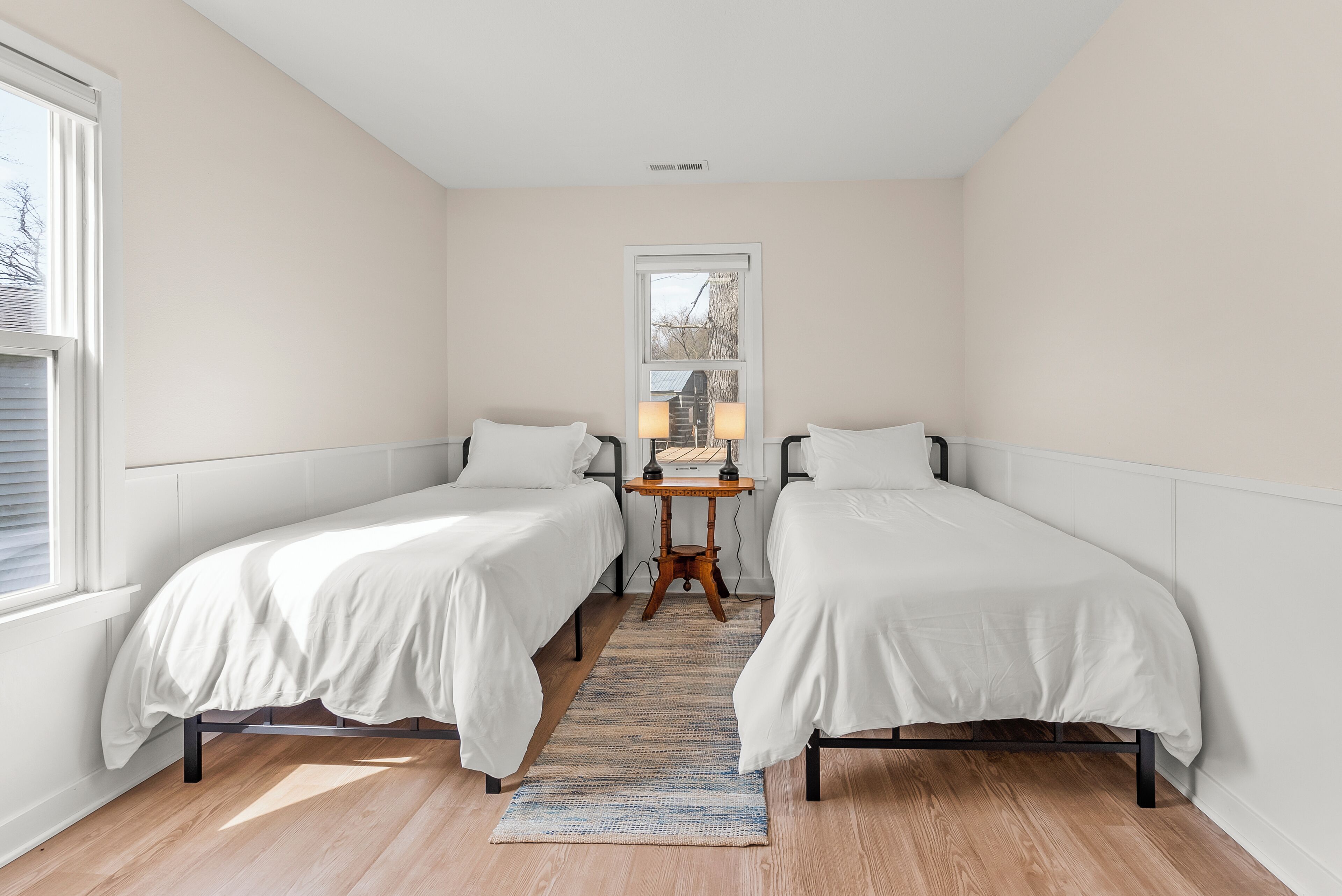 A bedroom with two single beds, a small table with lamps, and a window with blinds.