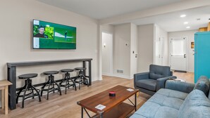 Una sala de estar moderna con un televisor de pantalla plana, un sofá gris, una mesa de centro de madera y una zona de bar con taburetes.