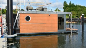A modern houseboat with a wooden deck, a circular window, and a metal railing.