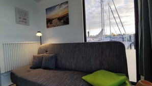 A room with a grey sofa, a green cushion, a lamp, a painting, and a view of boats through a window.